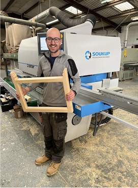 Man holding a wooden frame in front of a Soukup woodworking machine in a workshop