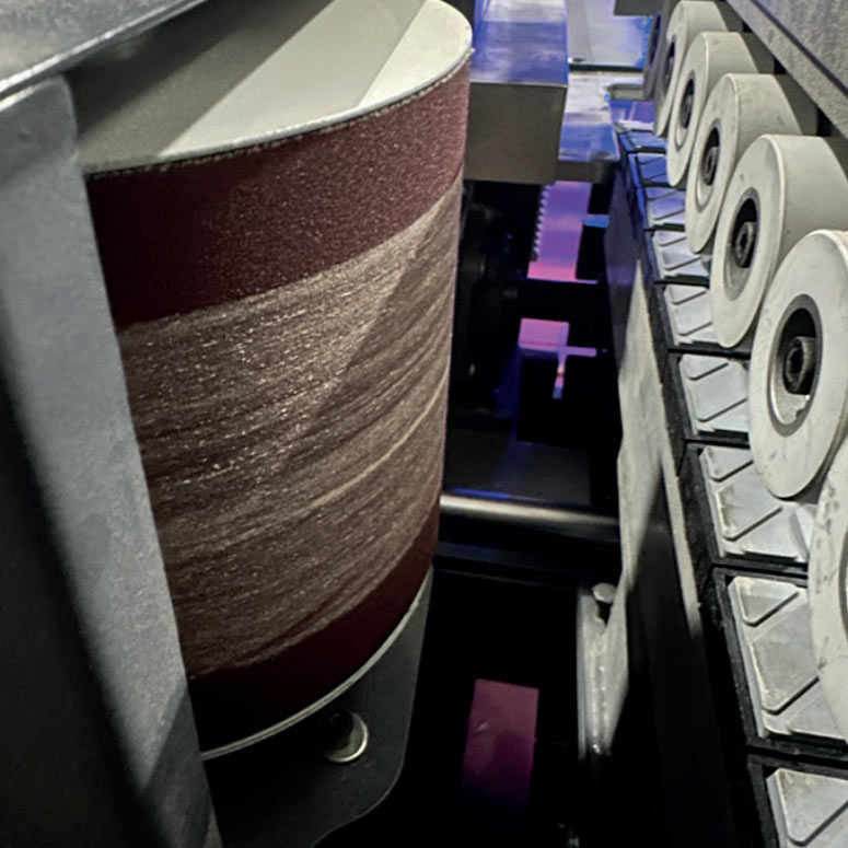 Close-up of a rotating abrasive drum sanding belt alongside a row of grinding wheels inside an industrial sanding machine