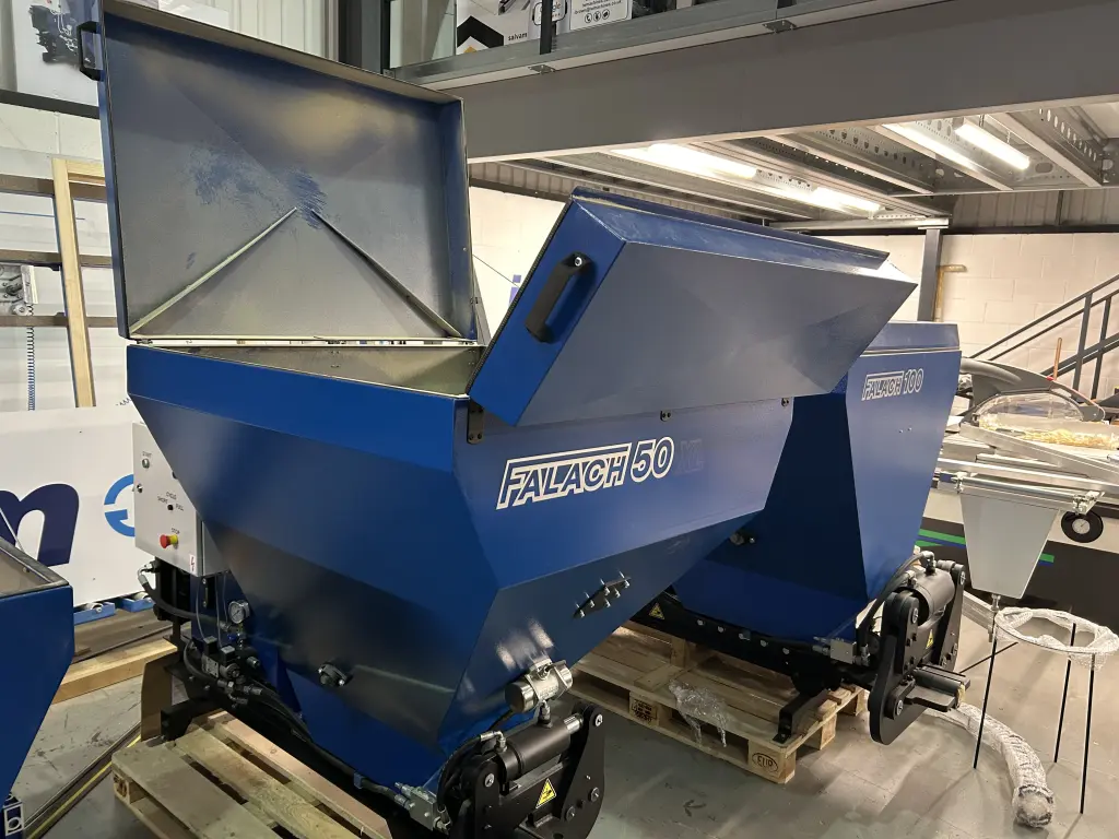 Blue Falach 50 briquette machine with open lid on a pallet inside an industrial workshop, with a Falach 100 visible behind it