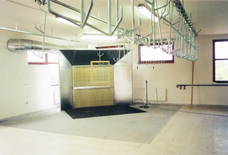 Industrial spray booth with overhead conveyor hooks, ventilation ducting, and a perforated yellow filter panel in a workshop