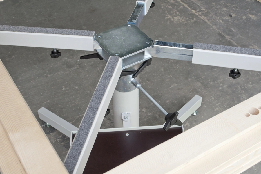 Underside of a white metal table base assembly with a central steel column, cross-frame arms, and adjustable feet on a concrete floor
