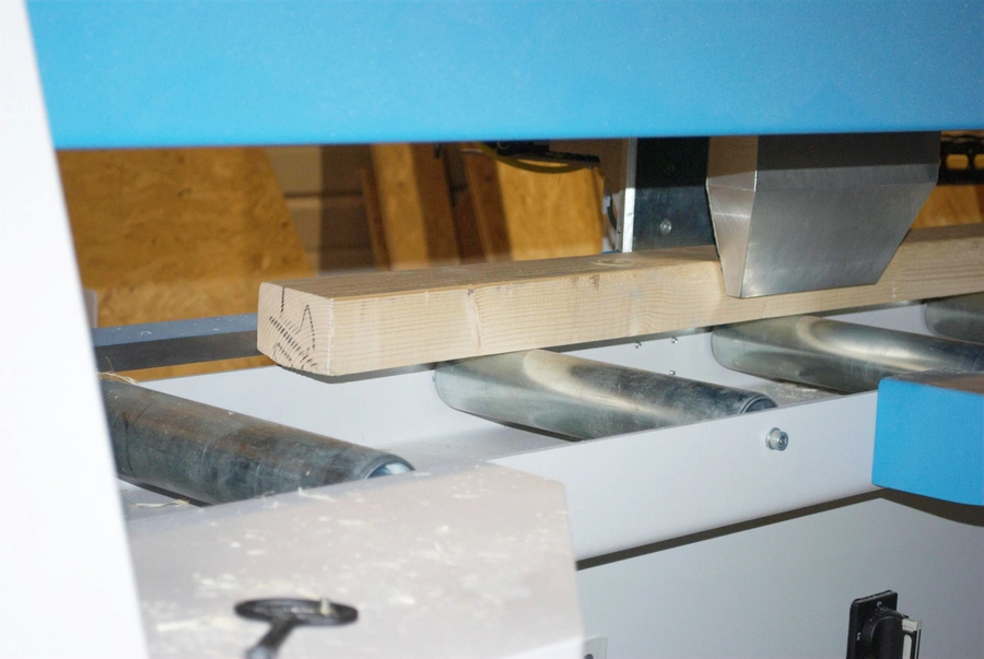 A timber plank being fed through a blue automatic cross cut saw machine on metal conveyor rollers