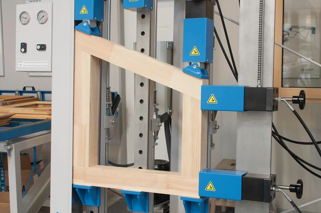 Wooden window frame held in a blue industrial press machine during manufacturing assembly