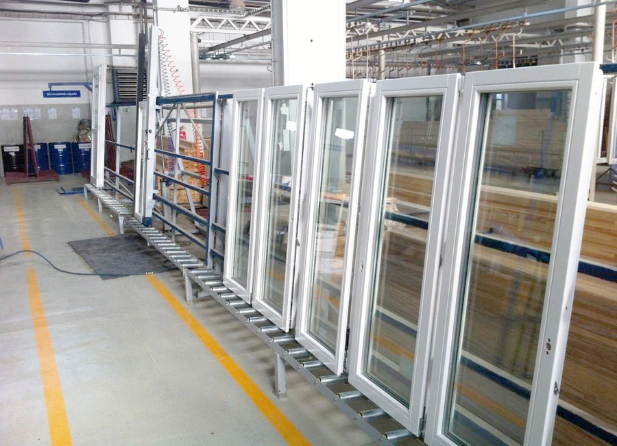Rows of white-framed glazed window units stored upright on a metal roller stand inside a large manufacturing facility