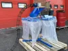 Blue IWM six bag carousel unit with top loading hopper, mounted on a wooden pallet outside a red industrial building.