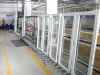 Rows of white-framed glazed window units stored upright on a metal roller stand inside a large manufacturing facility