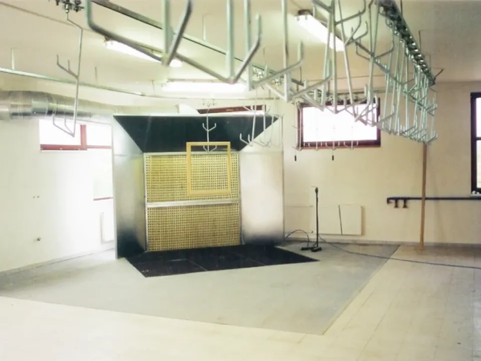 Industrial spray booth with overhead conveyor hooks, ventilation ducting, and a perforated yellow filter panel in a workshop